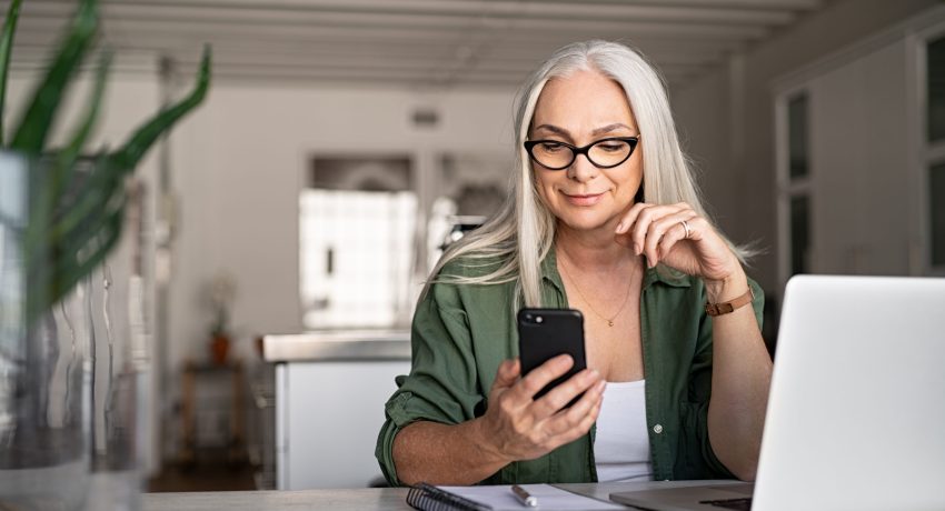 Happy senior woman using mobile phone while working at home with laptop